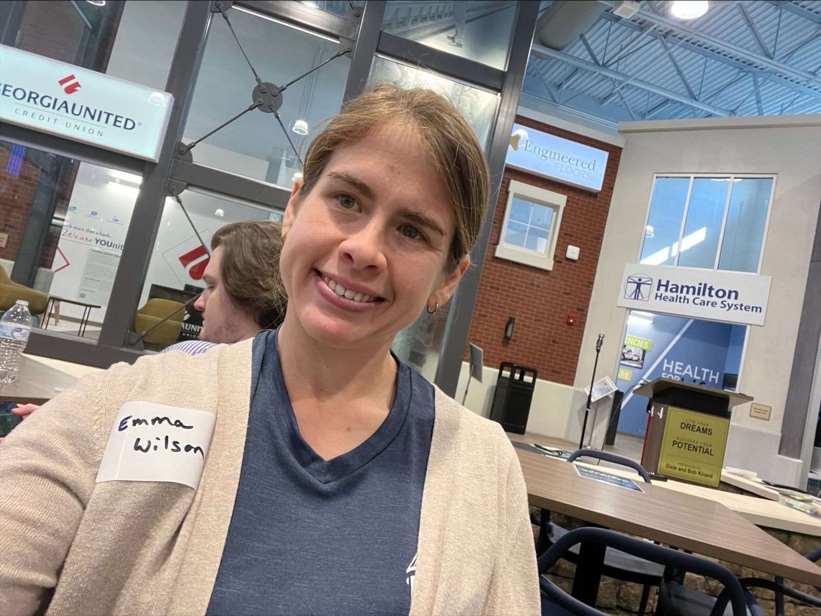 WSOB volunteer Emma Wilson taking a selfie in the JA Discover Center with a few of the simulator stores behind her, a GUCU bank and a Hamilton Health Care System building.