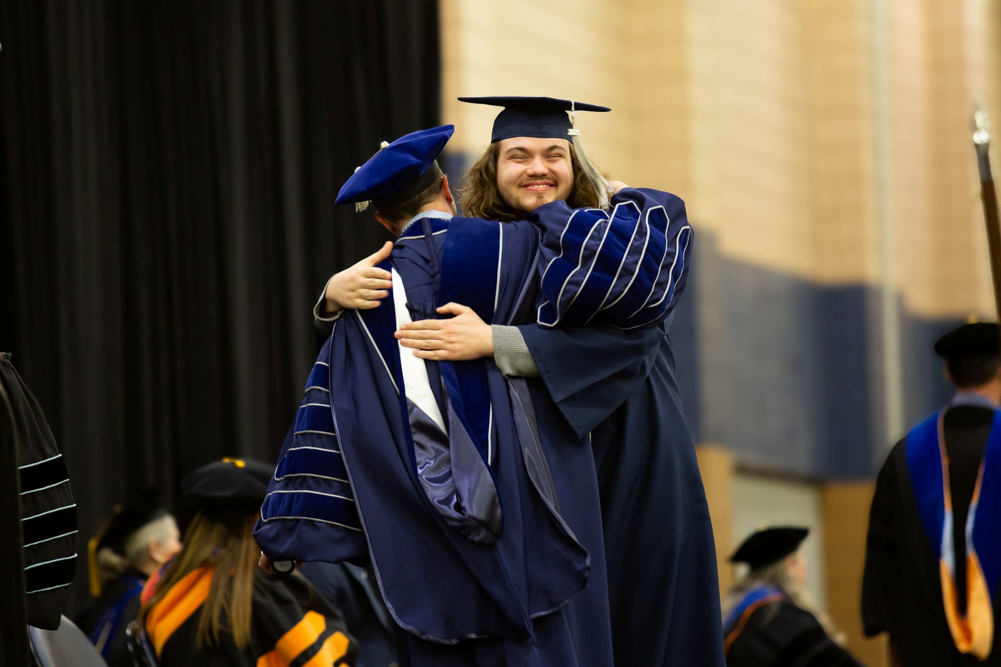 A college graduate embraces the college president for a hug on the commencement stage during graduation.