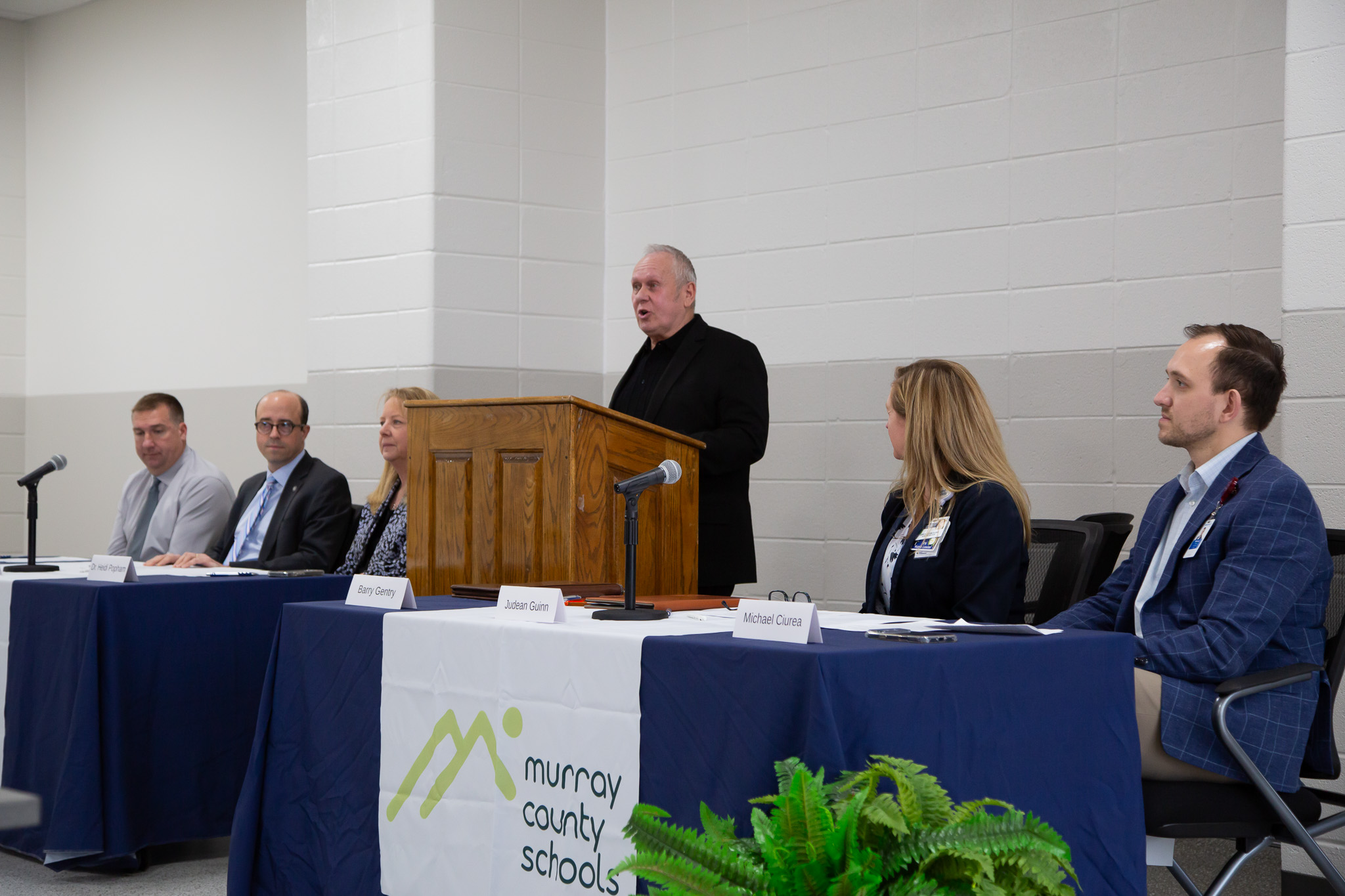 Four local leaders hold are seated behind a table listening to a man who is standing in the middle behind a podium speaks.