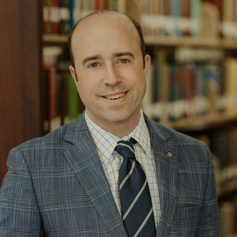 Photo of President Fuchko in a blue suit standing in front of bookshelves